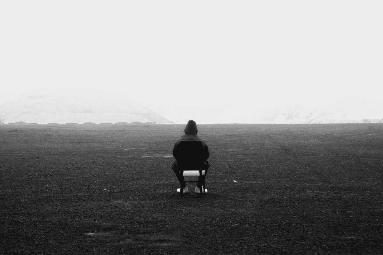 A man sits alone on a chair in an empty black landscape, facing away towards distant snow.